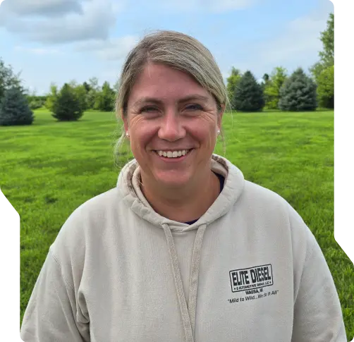 A smiling woman with blonde hair pulled back, wearing a light hoodie that says ELITE DIESEL, stands outdoors on green grass—highlighting the trusted work of certified diesel mechanics in Wausa NE—with trees and a cloudy sky in the background.