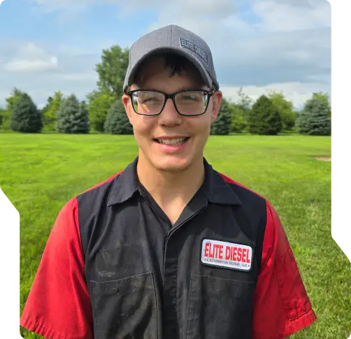 A young man wearing glasses, a gray Elite Diesel cap, and a black and red Elite Diesel work shirt stands smiling outdoors—proud to represent our team of certified diesel mechanics Wausa NE—on a green lawn with trees and a cloudy sky in the background.