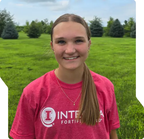 A young woman with straight brown hair in a low ponytail smiles outdoors. She is wearing a pink INTEGRA FORTIFIED T-shirt, standing on green grass, representing the experienced auto repair technicians Wausa trusts, with trees and a cloudy sky behind her.